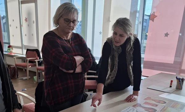 Zwei Frauen stehen vor einem Tisch und schauen sich Fotos an die verteilt sind, während eine Frau auf eines der Bilder zeigt.