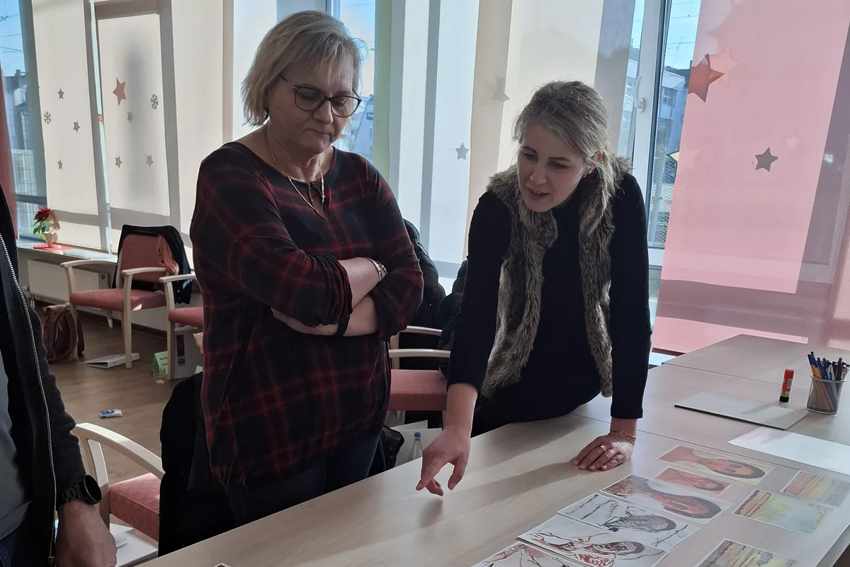 Zwei Frauen stehen vor einem Tisch und schauen sich Fotos an die verteilt sind, während eine Frau auf eines der Bilder zeigt.
