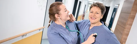 Das Bild zeigt zwei Frauen die einen blauen Überzieher anhaben und sich im Arm halten.Das Bild verlinkt zum Arbeitgeber-Video der Gesundheit