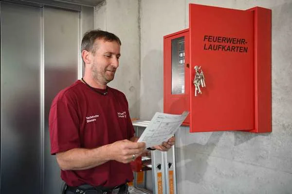 Das Bild zeigt einen jungen Mann der vor einem Stromkasten steht und ein rotes Poloshirt anhat.