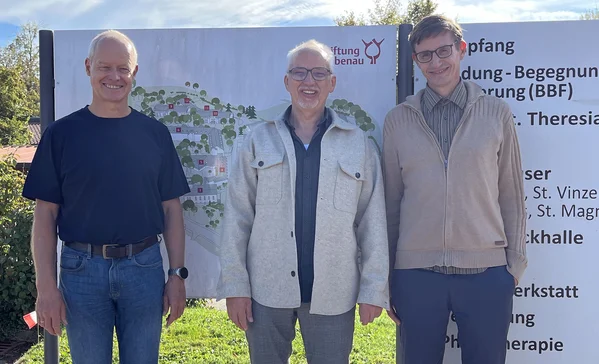 Drei lächelnde Männer stehen nebeneinander vor einem großen Informationsschild im Freien, auf einer Grasfläche, in legerer Kleidung.