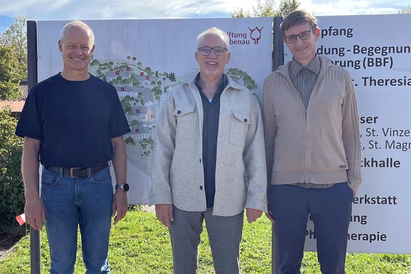 Drei lächelnde Männer stehen nebeneinander vor einem großen Informationsschild im Freien, auf einer Grasfläche, in legerer Kleidung.