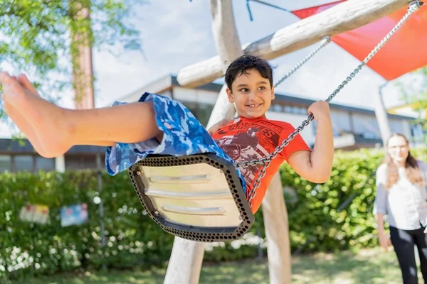 Das Bild zeigt einen kleinen Jungen der freudig schaukelt unter Aufsicht seiner Betreuerin.