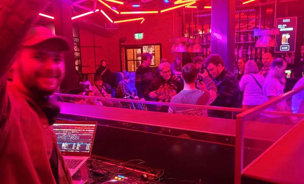 Ein DJ steht am DJ-Pult und Personen im Hintergrund feiern.