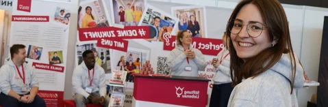 Eine Frau steht vor einem Messestand und schaut in die Kamera.