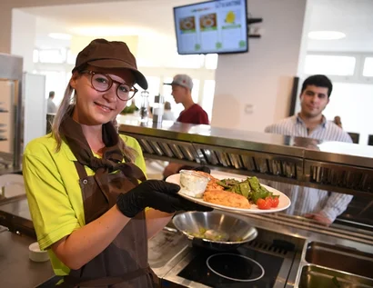 Das Bild zeigt eine Frau mit einem Teller in der Hand hinter einem Kantinentresen