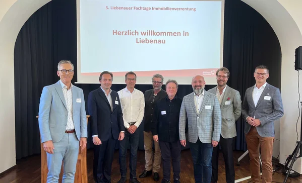 Acht Männer in Businesskleidung stehen lächelnd vor einer Projektionsleinwand mit der Aufschrift Herzlich willkommen in Liebenau bei einer Fachveranstaltung
