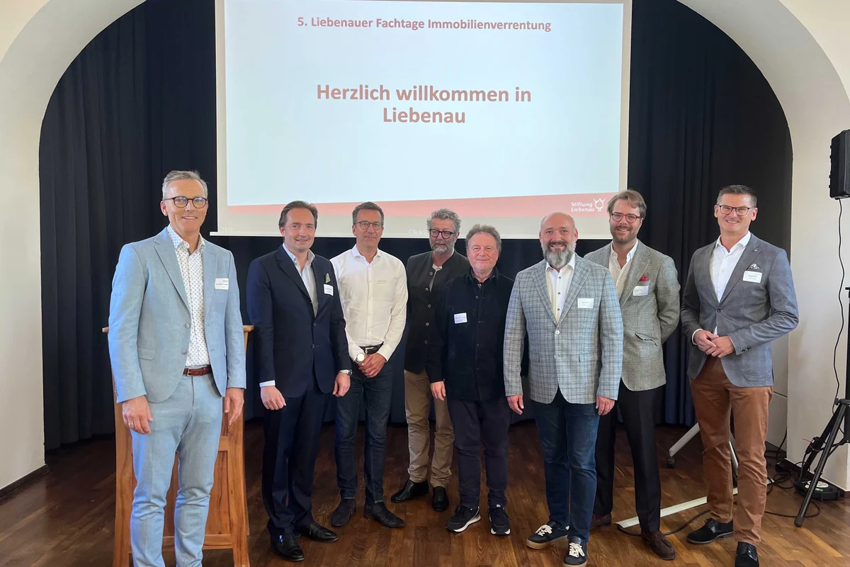 20 Jahre Immobilienverrentung in Deutschland Acht Männer in Businesskleidung stehen lächelnd vor einer Projektionsleinwand mit der Aufschrift Herzlich willkommen in Liebenau bei einer Fachveranstaltung