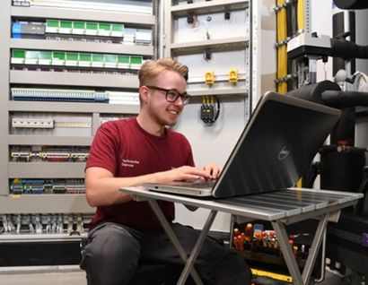 Das Bild zeigt einen jungen Mann mit Laptop an einer Schaltzentrale