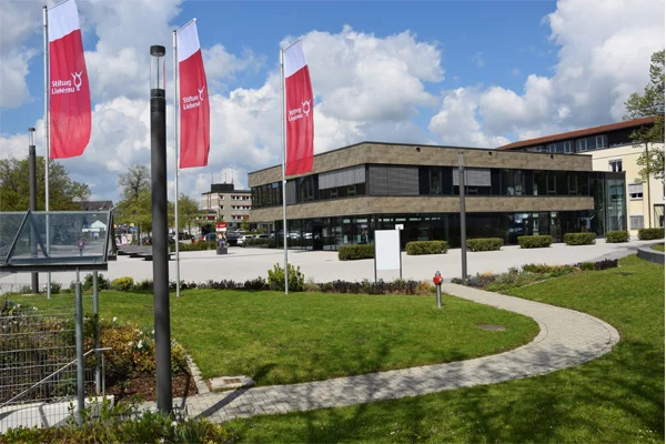 das foto zeigt den eingangsbereich der stiftung liebenau mit drei fahnen und wiese im vordergrund.