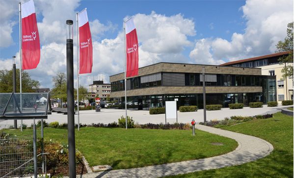 das foto zeigt den eingangsbereich der stiftung liebenau mit drei fahnen und wiese im vordergrund.