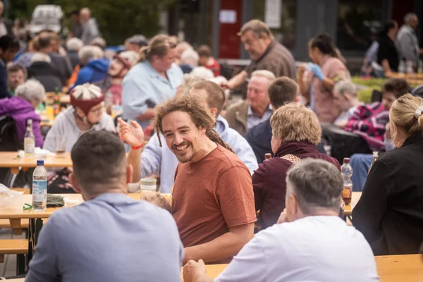 Auf dem Foto sind mehrere Personen auf Bierbänken zu sehen. In der Mitte sitzt ein Mann der lächelt.