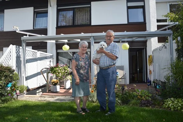 Das Bild zeigt ein Ehepaar, das gemeinsam in ihrem Garten steht. Die Frau hält einen kleinen weißen Hund in der Hand und sie schauen gemeinsam in die Kamera