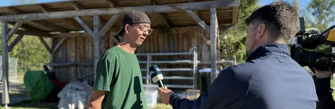 Ein Mann in grünem T-Shirt und Baseballcap wird im Freien von einem Reporter mit Mikrofon und einem Kameramann vor einem hölzernen Unterstand interviewt.