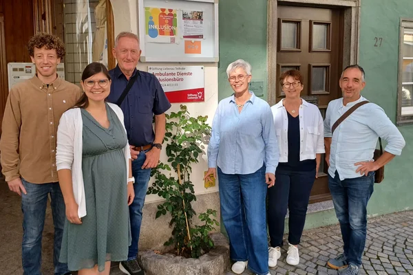 Sechs lächelnde Erwachsene unterschiedlichen Alters stehen nebeneinander vor einem Haus- bzw. Büroeingang mit Pflanzen und einem Schild mit den Worten "Inklusion" und "Ambulante Dienste