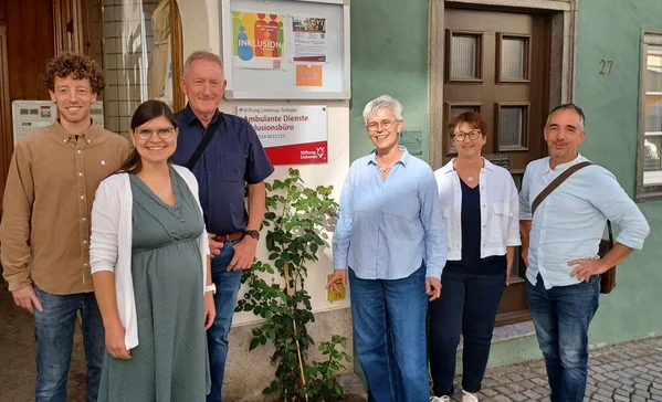 Sechs lächelnde Erwachsene unterschiedlichen Alters stehen nebeneinander vor einem Haus- bzw. Büroeingang mit Pflanzen und einem Schild mit den Worten "Inklusion" und "Ambulante Dienste