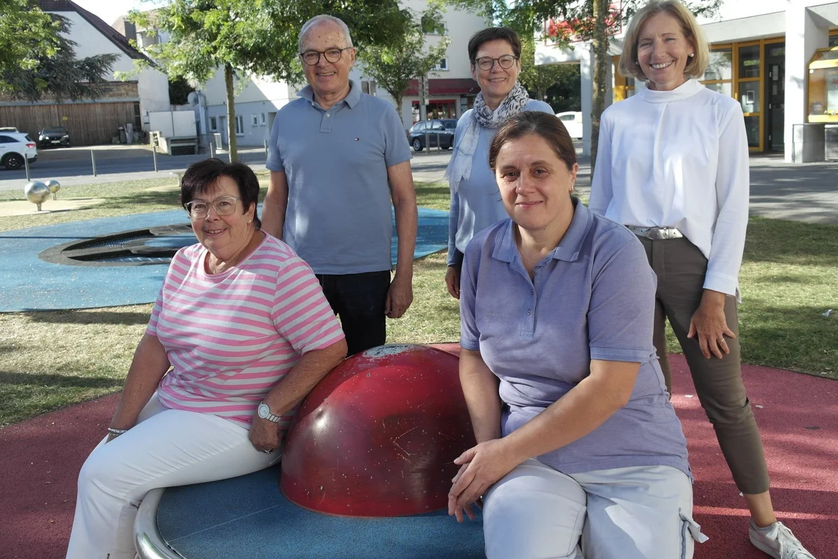Gruppenbild außen Fünf Erwachsene auf einem Spielplatz im Freien: drei Frauen sitzen um eine rote Kugel, zwei Personen stehen dahinter. Alle blicken lächelnd zur Kamera.