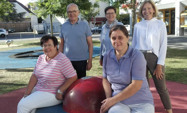 Fünf Erwachsene auf einem Spielplatz im Freien: drei Frauen sitzen um eine rote Kugel, zwei Personen stehen dahinter. Alle blicken lächelnd zur Kamera.