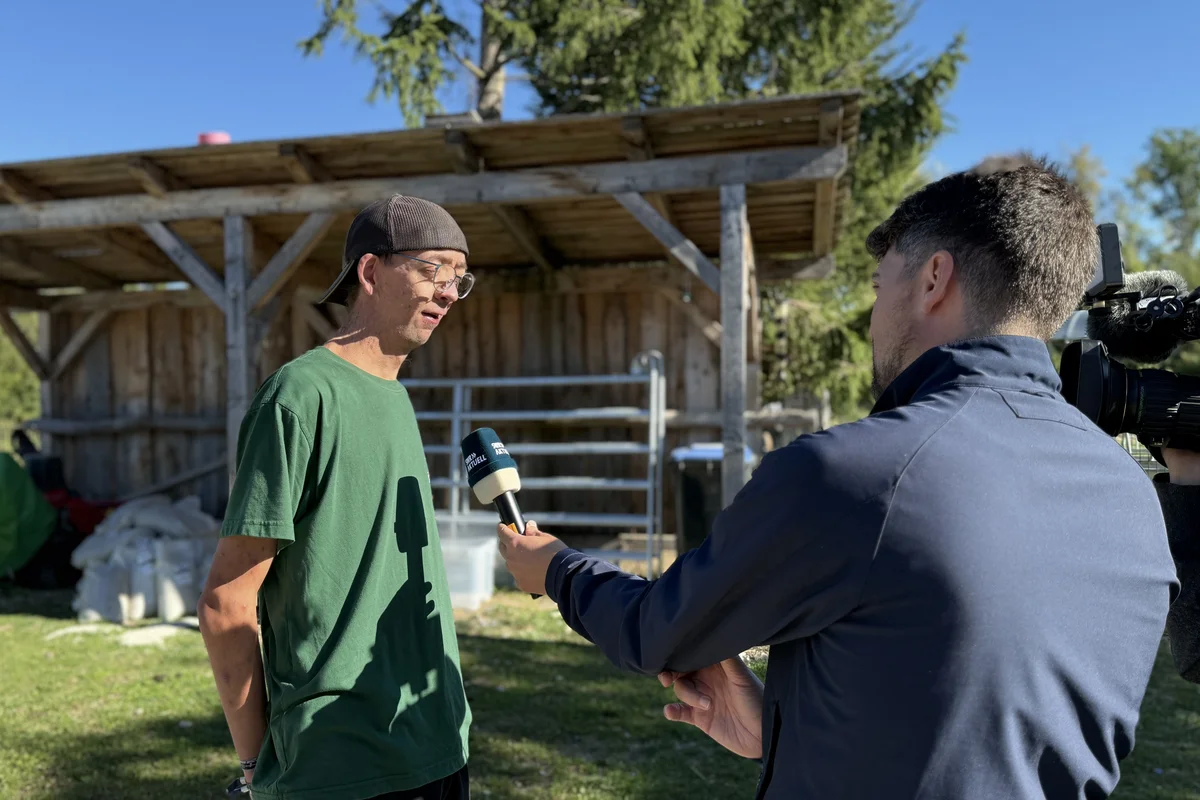 Stöckle Ein Mann in grünem T‑Shirt wird im Freien von einem Reporter mit Mikrofon interviewt, ein Kameramann filmt die Szene vor einem Holzunterstand.
