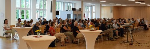 Das Bild zeigt einen Saal mit sitzenden Zuhörern und drei Tischen im Vordergrund