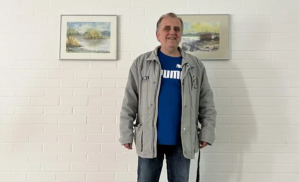 Ein Mann steht vor einer weißen Wand mit Kunstgemälde im Hintergrund und lächelt in die Kamera.