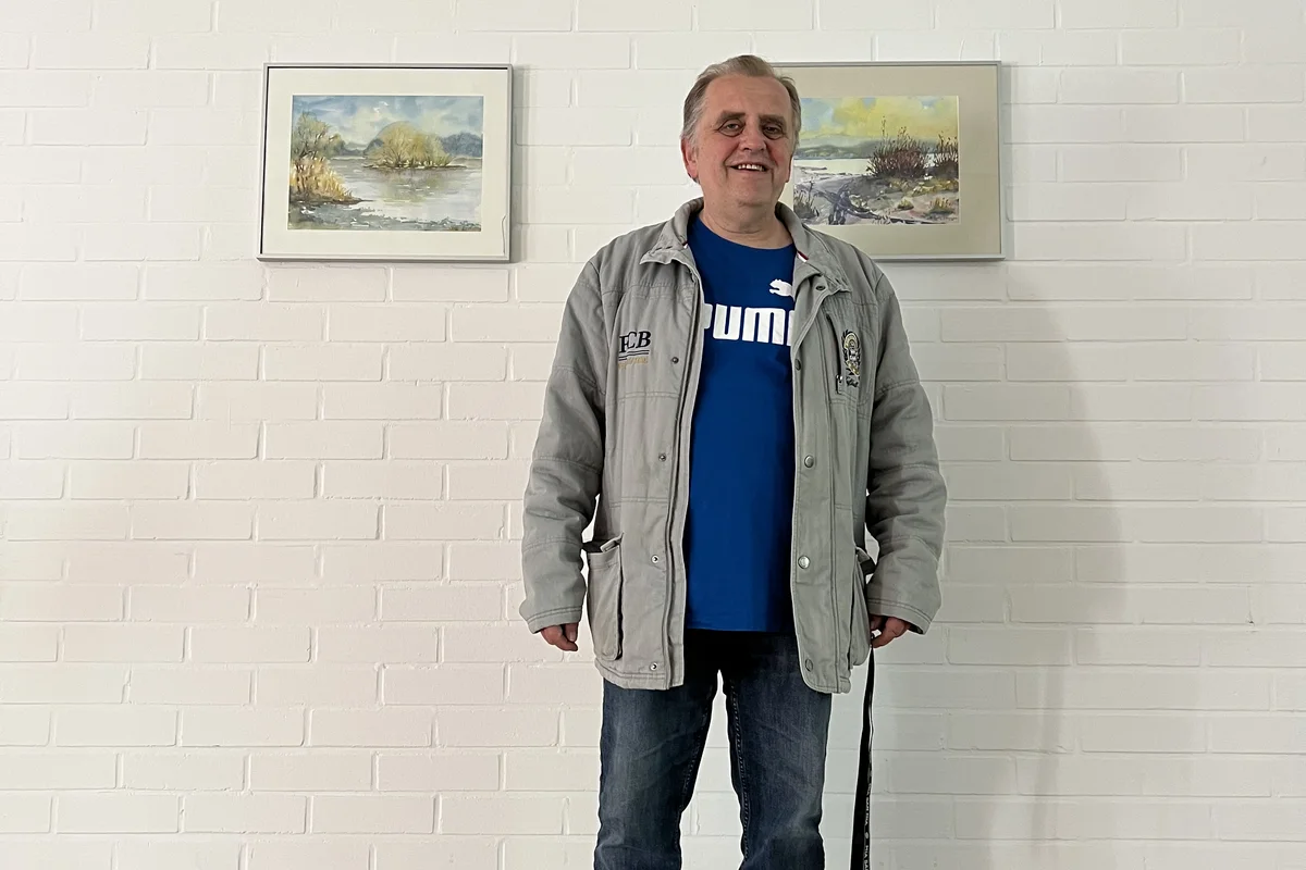 Thomas Tischmacher Ein Mann steht vor einer weißen Wand mit Kunstgemälde im Hintergrund und lächelt in die Kamera.