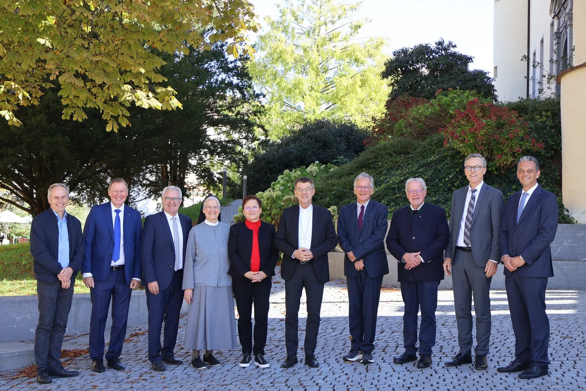 Bischof Krämer besucht Stiftung Liebenau Gruppenbild Elf Personen unterschiedlicher Altersgruppen in formeller Kleidung stehen nebeneinander im Freien vor Büschen und einem Gebäude, lächelnd und für ein Gruppenfoto posierend.