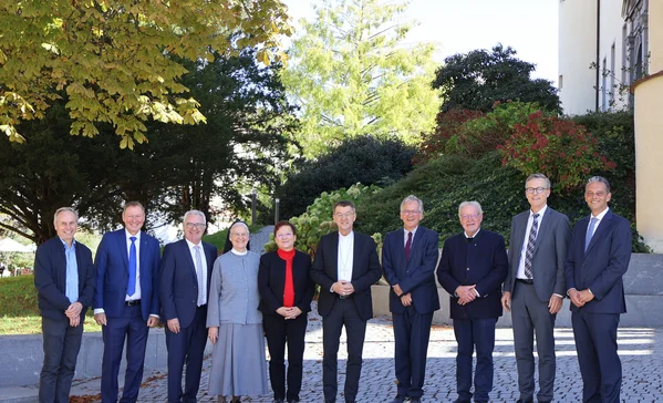 Bischof Krämer besucht Stiftung Liebenau Gruppenbild Elf Personen unterschiedlicher Altersgruppen in formeller Kleidung stehen nebeneinander im Freien vor Büschen und einem Gebäude, lächelnd und für ein Gruppenfoto posierend.