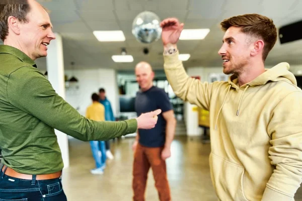 Schwierige Situationen im Berufsalltag frühzeitig erkennen und deeskalieren Das Bild zeigt zwei Männer die sich gerade die Han geben. Der eine hat einen Blauen Anzug an und der andere einen gelben Pullover