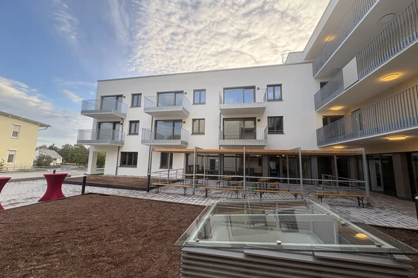 Innenhof eines modernen weißen Mehrfamilienhauses mit Balkonen, Außenterrasse mit Sitzbänken, barrierefreier Rampe und Glas-Lichtschacht im Vordergrund unter bewölktem Himmel