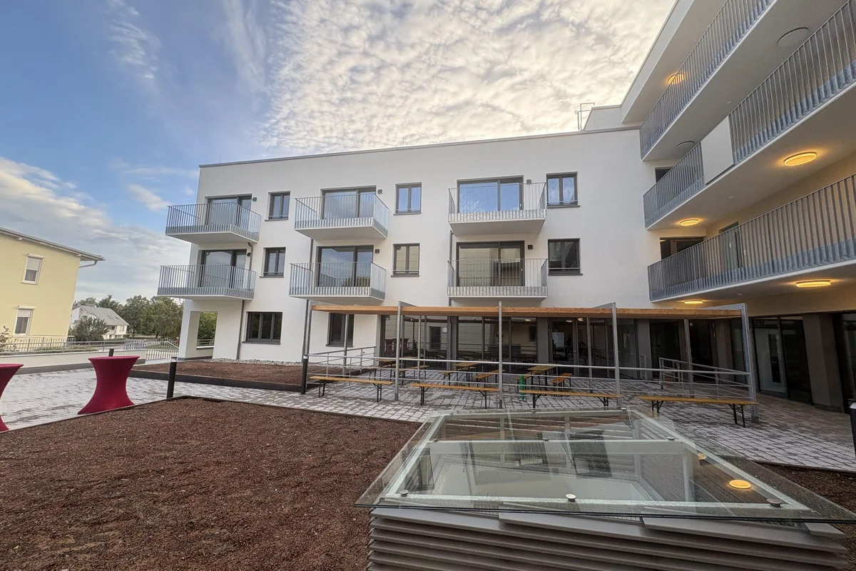 Tag der offenen Baustelle Innenhof eines modernen weißen Mehrfamilienhauses mit Balkonen, Außenterrasse mit Sitzbänken, barrierefreier Rampe und Glas-Lichtschacht im Vordergrund unter bewölktem Himmel