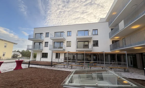 Innenhof eines modernen weißen Mehrfamilienhauses mit Balkonen, Außenterrasse mit Sitzbänken, barrierefreier Rampe und Glas-Lichtschacht im Vordergrund unter bewölktem Himmel