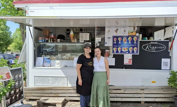 Zwei Frauen stehend lächelnd vor einem großen Eiswagen.
