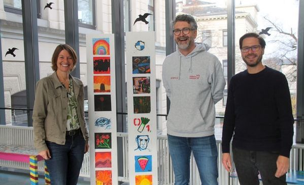 Das Bild zeigt drei personen die vor der Kamera stehenund lachen. Zwischen ihnen steht eine Säule an der Plakate angebracht sind