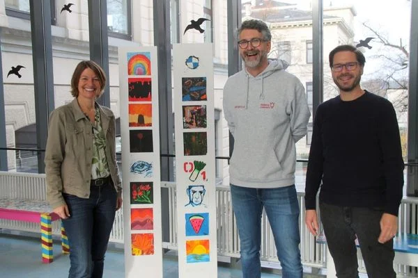 Das Bild zeigt drei personen die vor der Kamera stehenund lachen. Zwischen ihnen steht eine Säule an der Plakate angebracht sind