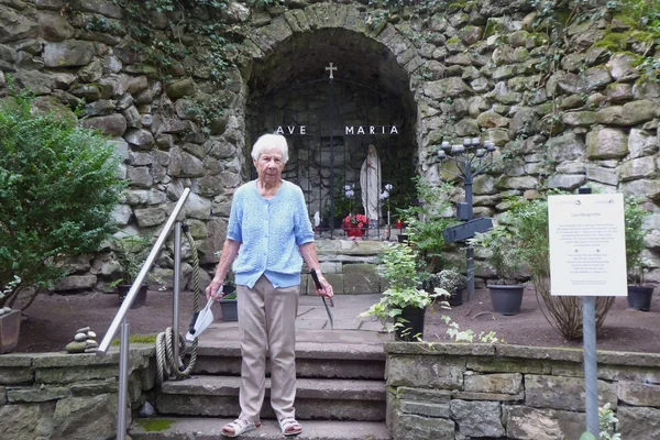 Ältere Frau steht auf einer Treppe vor einer steinernen Mariengrotte mit Marienstatue, Kreuz und dem Schriftzug "Ave Maria