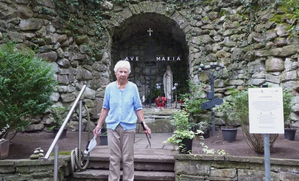Ältere Frau steht auf einer Treppe vor einer steinernen Mariengrotte mit Marienstatue, Kreuz und dem Schriftzug "Ave Maria