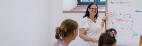 Eine Frau an einem Whiteboard erklärt etwas und lächelt in die Kamera.