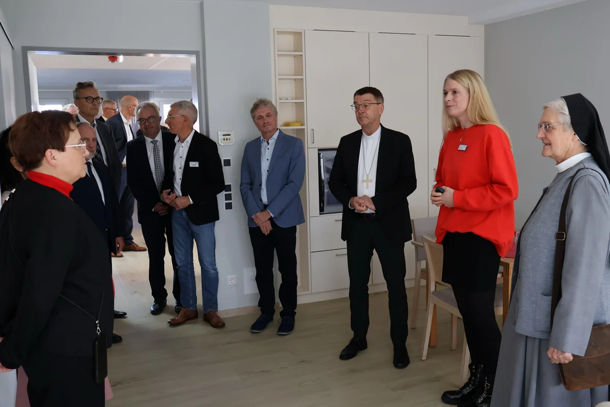 Bischof Krämer besucht Stiftung Liebenau Hospiz Mehrere Erwachsene stehen in einem hellen Zimmer; einige tragen formelle Kleidung, darunter ein Mann mit Priesterkragen und eine Nonne. Die Gruppe hört einer Frau zu, die spricht.