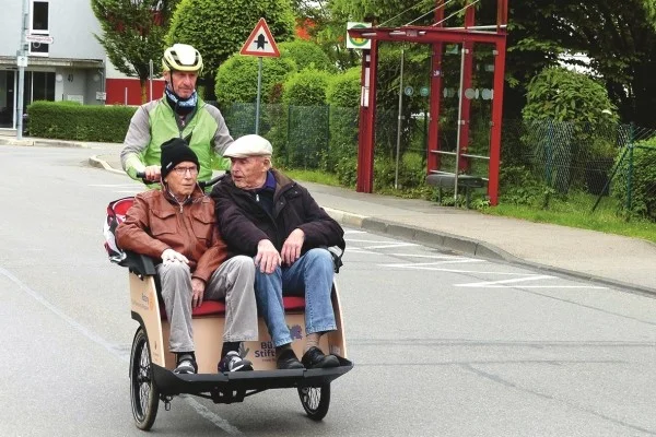 Das Bild zeigt einen Mann der mit zwei älteren Personen Rikscha fährt