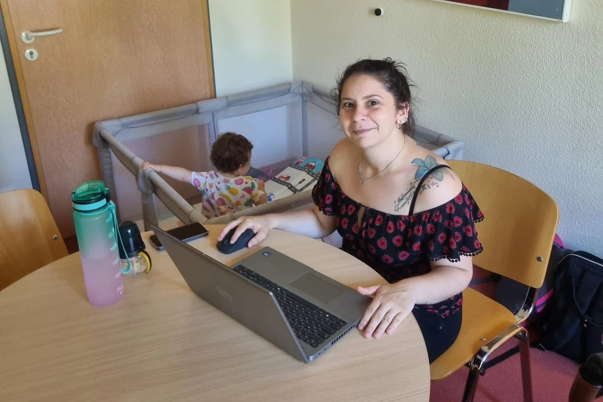 Familienfreundliche Arbeitsgestaltung Eine Frau sitzt am Tisch und lächelt in die Kamera, während im Hintergrund ein Baby in seinem Laufgitter spielt.