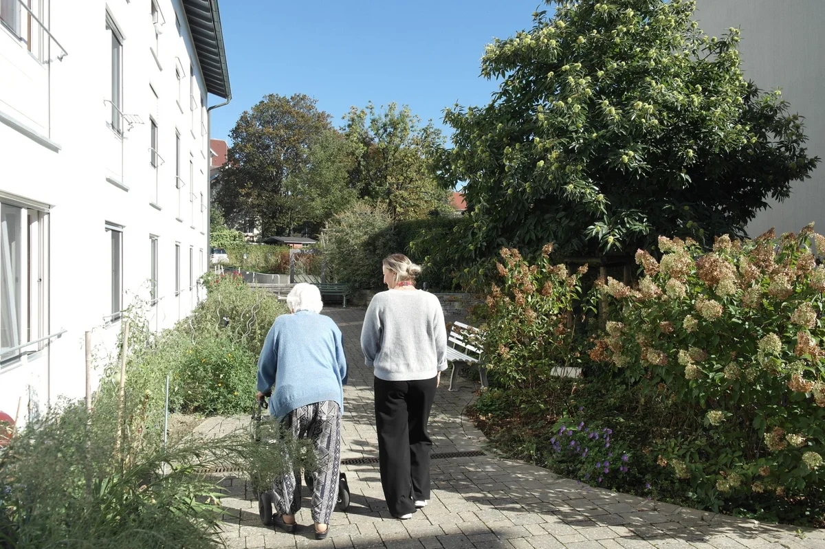 Demenzgarten Zwei Frauen gehen auf einem sonnigen Gartenweg zwischen Wohngebäuden; eine nutzt einen Rollator, die andere begleitet sie.