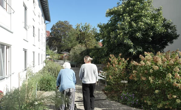 Zwei Frauen gehen auf einem sonnigen Gartenweg zwischen Wohngebäuden; eine nutzt einen Rollator, die andere begleitet sie.