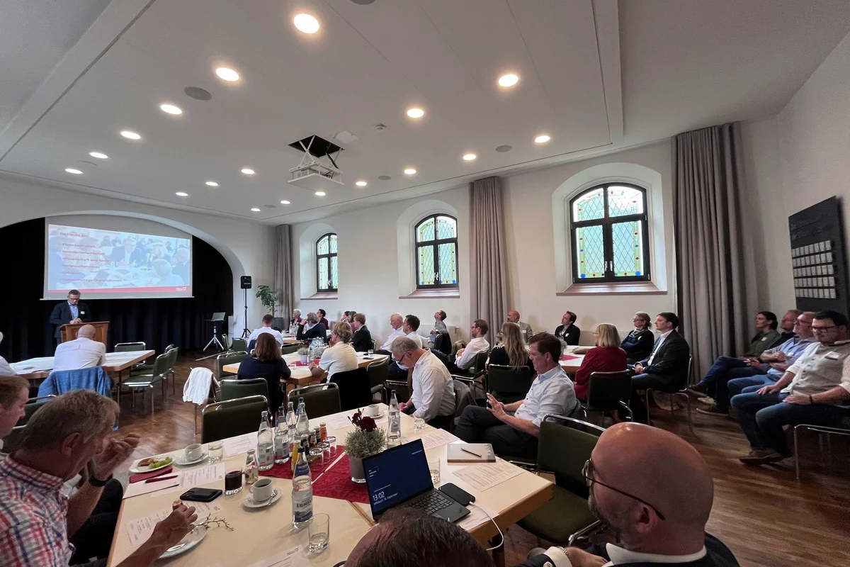 20 Jahre Immobilienverrentung in Deutschland Konferenzraum mit mehreren Tischen, Teilnehmern, die einem Vortrag an einem Projektorbildschirm zuschauen; informelle Besprechungsszene mit Notebooks, Getränken und Blick auf Präsentierenden.