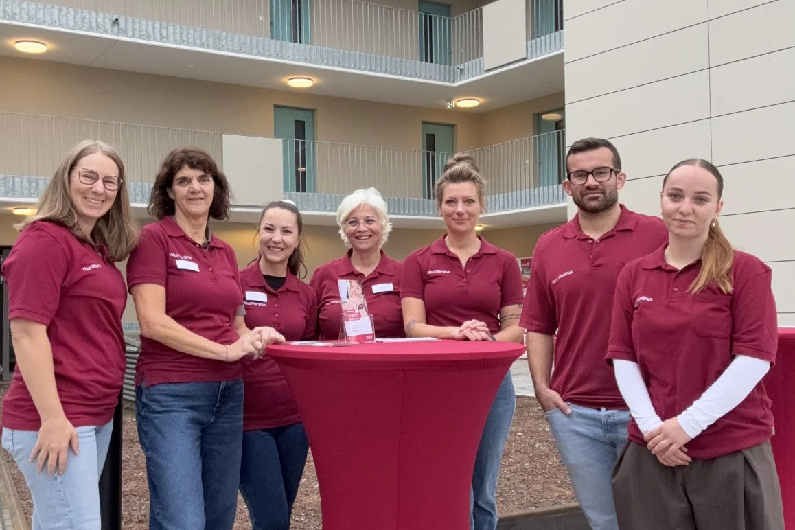 Tag der offenen Baustelle Sieben Personen in einheitlichen dunkelroten Poloshirts stehen lächelnd um einen hohen, rot bezogenen Stehtisch in einem hellen Atrium.