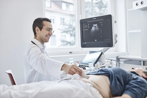 Das Bild zeigt einen Doktor der ein Ultraschall an einer Dame durchführt die auf einer Liege liegt.