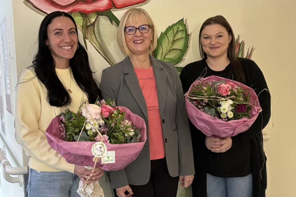 Team Drei Frauen lachen in die Kamera und halten Blumensträuße.