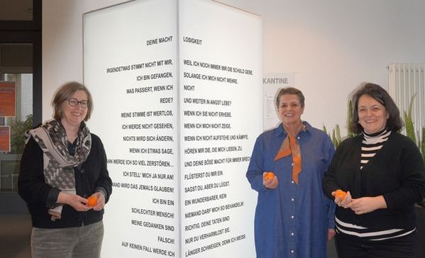 Das Bild zeigt drei Frauen, die gesammelt vor einer Lichtersäule stehen. Sie halten Orangen in der Hand und schauen in die Kamera