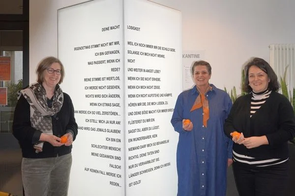 Das Bild zeigt drei Frauen, die gesammelt vor einer Lichtersäule stehen. Sie halten Orangen in der Hand und schauen in die Kamera