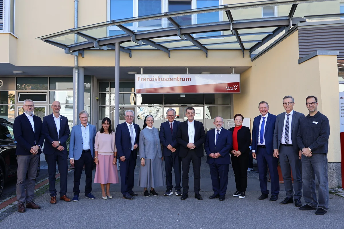 Bischof Krämer besucht Stiftung Liebenau Friedrichshafen Gruppe von dreizehn Personen in formeller Kleidung, die vor dem Eingang eines Gebäudes mit dem Schild "Franziskuszentrum" stehen und in die Kamera lächeln.